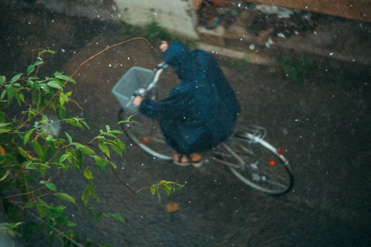 Person cycling in rain wearing a raincoat, captured from above, showcasing motion and weather.