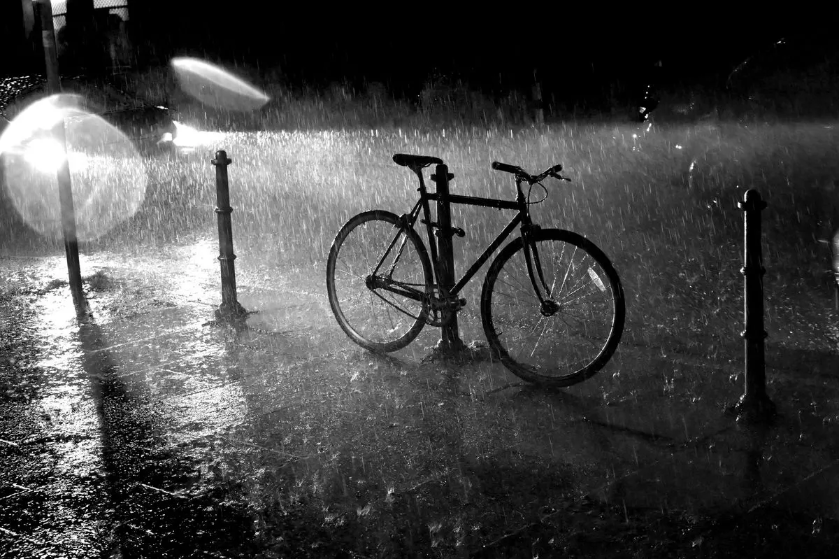 A solitary bicycle parked in the rain on an urban street at night, illuminated by streetlights.