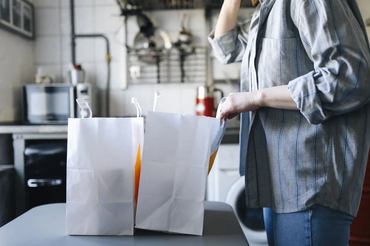 Woman in casual attire handling paper bags for delivery service in a kitchen setting.