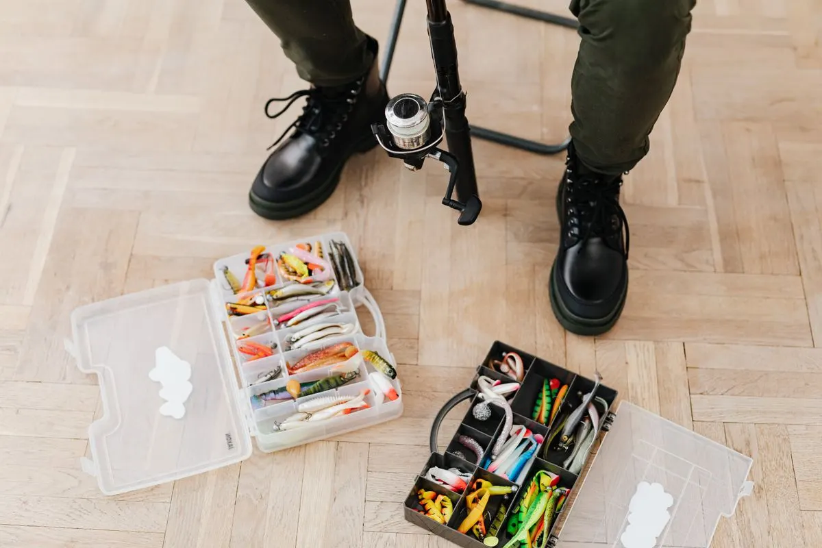 Fishing tackle boxes with lures and equipment beside boots on a wooden floor.