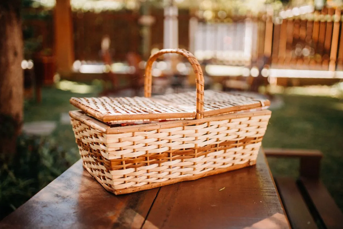 Wicker basket for picnic with lid placed on table in backyard with green lawn and wooden fence on blurred background